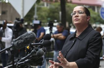 A woman speaking at a press conference, gesturing with her hand, surrounded by cameras and reporters.
