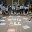 A group of protesters holding signs and celebrating LGBTQ rights gathered outdoors, with colorful chalk messages on the ground. A group of protesters holding signs and celebrating LGBTQ rights gathered outdoors, with colorful chalk messages on the ground.