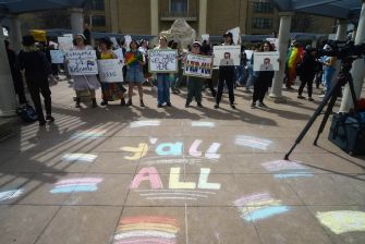 A group of protesters holding signs and celebrating LGBTQ rights gathered outdoors, with colorful chalk messages on the ground.