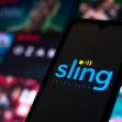 A smartphone displaying the Sling Television logo, with a blurred background of various streaming content. A smartphone displaying the Sling Television logo, with a blurred background of various streaming content.