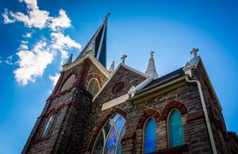 A perspective view of a stone church with a tall spire against a blue sky with clouds.