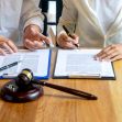 Two individuals discussing and signing contracts at a table, with a gavel in the foreground. Two individuals discussing and signing contracts at a table, with a gavel in the foreground.