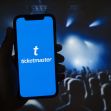 A person holding a smartphone displaying the Ticketmaster logo in front of a concert crowd. A person holding a smartphone displaying the Ticketmaster logo in front of a concert crowd.