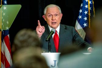 Former Attorney General Jeff Sessions speaking at a podium with American flags in the background.