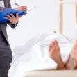 A medical professional in a suit observes a covered body while taking notes. A medical professional in a suit observes a covered body while taking notes.