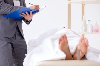 A medical professional in a suit observes a covered body while taking notes.