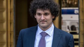 A man in a suit walks outside a building, amidst a backdrop of blurred vehicles and urban scenery.