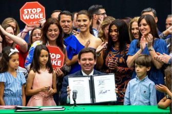 Governor Ron DeSantis holding a signed document promoting the "Stop WOKE Act" surrounded by supporters, including children and individuals holding "Stop WOKE" signs.