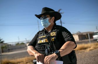 A law enforcement officer in a cowboy hat and face mask stands outside, overseeing a situation related to evictions.