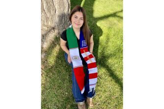 A young woman sitting on grass with a graduation sash featuring the Mexican flag on one side and the American flag on the other, smiling at the camera.
