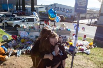 Two teenagers embrace in front of a memorial at Saugus High School, surrounded by flowers and balloons, honoring victims of a school shooting.
