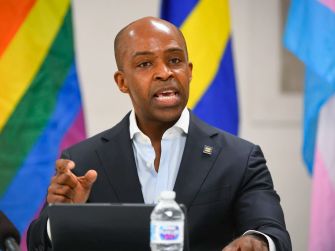 Alphonso David speaking at a press conference, with rainbow flags in the background.