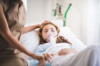 A young girl in a hospital bed receiving medical care with an oxygen mask, indicating a serious health condition.