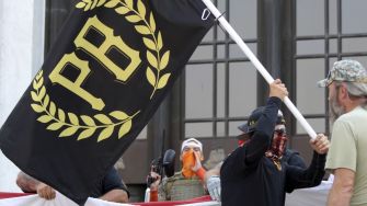 A group of individuals at a protest holding a black flag with the initials "PB" representing the Proud Boys organization.