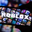 A hand holding a smartphone displaying the Roblox logo, with various game images in the background. A hand holding a smartphone displaying the Roblox logo, with various game images in the background.