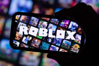 A hand holding a smartphone displaying the Roblox logo, with various game images in the background.
