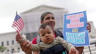 A woman holding a baby and a sign that reads "AMERICAN BORN CHILDREN ARE AMERICAN CHILDREN," with a small American flag, outside a courthouse.