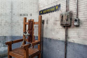 Electric chair in a prison setting, labeled "BLOCK D" and "HIGH VOLTAGE."