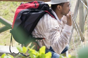 A person sitting on a fence with a backpack, looking contemplative, possibly reflecting on their journey or the challenges faced by migrants.