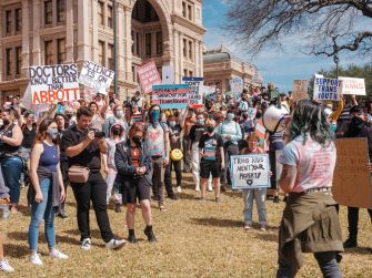 Demonstrators gather outside a building holding signs in support of transgender rights and against Texas Governor Abbott's order regarding gender-affirming care for minors.
