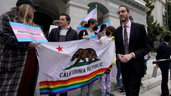 Demonstrators holding signs and a California flag with rainbow colors, advocating for transgender rights outside a government building.