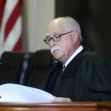 A judge in a courtroom reading documents, with an American flag in the background. A judge in a courtroom reading documents, with an American flag in the background.