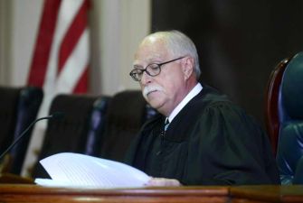 A judge in a courtroom reading documents, with an American flag in the background.