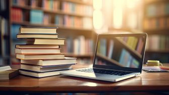 A stack of books next to a laptop on a desk in a library setting.
