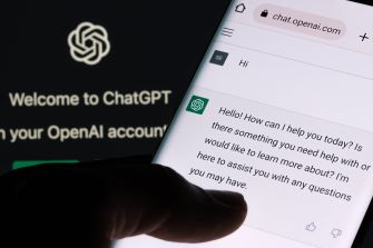 A hand holding a smartphone displaying the ChatGPT interface, with a greeting message on the screen and a dark background featuring the ChatGPT logo.