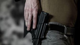 A close-up image of a person holding a handgun in a holster, resting on their hip. The background is blurred, focusing on the firearm.