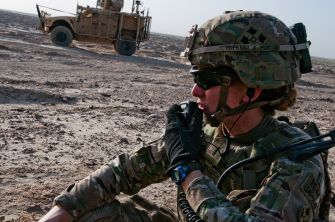 A soldier communicating via radio in a military setting, with a vehicle in the background.