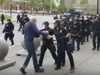 Two police officers pushing an elderly man during a protest, with a line of police in riot gear in the background.