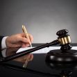 A judge's gavel sits on a desk beside a hand holding a pen writing on a document, symbolizing court proceedings and legal decisions. A judge's gavel sits on a desk beside a hand holding a pen writing on a document, symbolizing court proceedings and legal decisions.