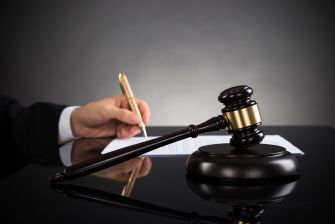 A judge's gavel sits on a desk beside a hand holding a pen writing on a document, symbolizing court proceedings and legal decisions.