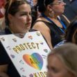 A person holding a sign that reads "PROTECT ALL KIDS" at a gathering, with a focus on LGBTQ+ rights and advocacy. A person holding a sign that reads "PROTECT ALL KIDS" at a gathering, with a focus on LGBTQ+ rights and advocacy.
