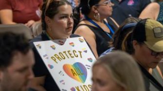 A person holding a sign that reads "PROTECT ALL KIDS" at a gathering, with a focus on LGBTQ+ rights and advocacy.