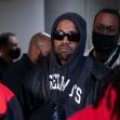 Kanye West wearing sunglasses and a black hoodie, surrounded by other individuals. Kanye West wearing sunglasses and a black hoodie, surrounded by other individuals.