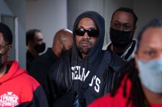 Kanye West wearing sunglasses and a black hoodie, surrounded by other individuals.