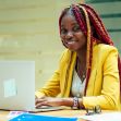 A smiling woman with braided hair sits at a desk with a laptop, wearing a yellow blazer, in a bright office setting. A smiling woman with braided hair sits at a desk with a laptop, wearing a yellow blazer, in a bright office setting.