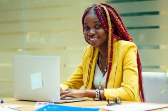 A smiling woman with braided hair sits at a desk with a laptop, wearing a yellow blazer, in a bright office setting.