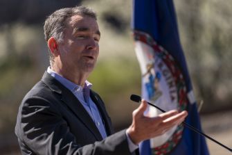 Virginia Governor Ralph Northam speaking during a press event, with the Virginia state flag in the background.