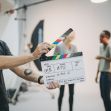 A person holding a clapperboard in a film audition setting with two actors in the background. A person holding a clapperboard in a film audition setting with two actors in the background.