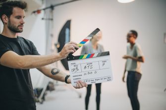 A person holding a clapperboard in a film audition setting with two actors in the background.