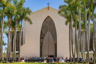 A modern church building with palm trees in the foreground, where a group of people is gathered.