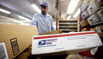 A postal worker handling packages inside a USPS facility, with a focus on a "Priority Mail" box in the foreground.