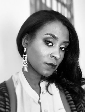 A black and white portrait of a woman with long hair, wearing statement earrings and a button-up shirt, looking thoughtfully at the camera.