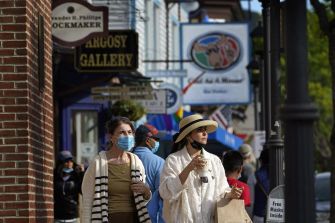 Two women wearing masks walk along a busy street with various shops and signage in the background.