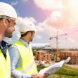 Two construction workers in hard hats and reflective vests discussing plans at a construction site with a crane in the background. Two construction workers in hard hats and reflective vests discussing plans at a construction site with a crane in the background.
