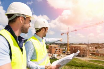 Two construction workers in hard hats and reflective vests discussing plans at a construction site with a crane in the background.