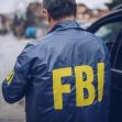 A person wearing a blue jacket with "FBI" printed in yellow on the back, standing on a street near a vehicle. A person wearing a blue jacket with "FBI" printed in yellow on the back, standing on a street near a vehicle.